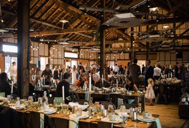 Wedding guests gathered for a barn reception dinner at Freight in Leadville, featuring long wooden dining tables set with greenery and candles under high ceilings with exposed wooden beams.